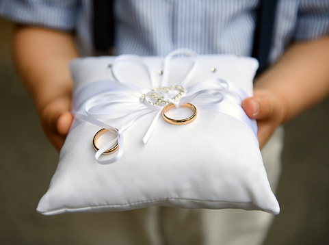 Pageboy holding two gold wedding bands or rings on a cushion decorated with white bow during the marriage ceremony at a church in close up