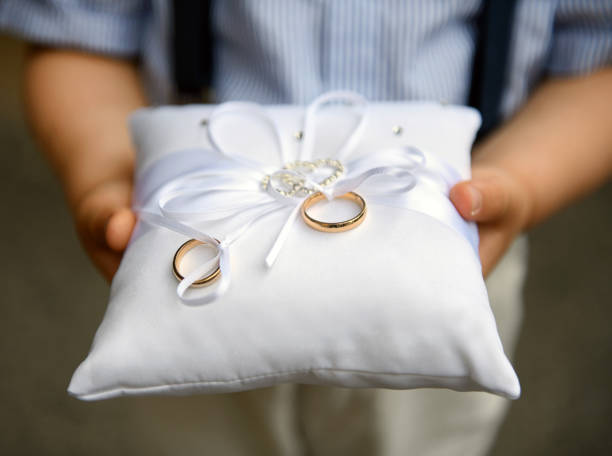 Kid holding two gold wedding bands on a cushion Pageboy holding two gold wedding bands or rings on a cushion decorated with white bow during the marriage ceremony at a church in close up ring bearer stock pictures, royalty-free photos & images