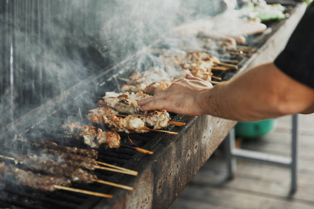 istock Hand of young man grilling some meat and vegetables on long rectangular barbecue with smoke on the street outdoor. Summer barbecue cooking. Barbecue with raw vegetables. BBQ on the terrace 1286166576