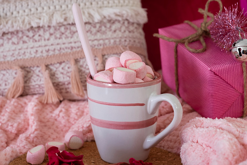 Close-up view on a ceramic cup with hot cocoa with marshmallow. Pink home corner with blanket, pillow and Christmas gift.  Holidays and happy people concept