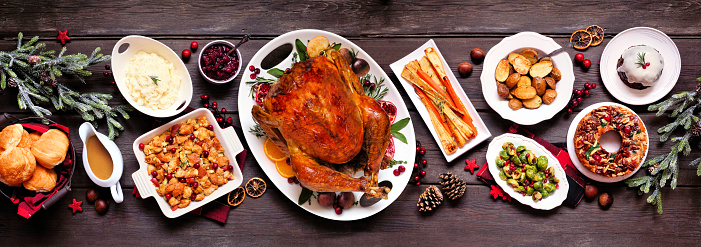 Traditional Christmas turkey dinner. Top view panoramic table scene on a dark wood banner background. Turkey, potatoes and sides, dressing, fruit cake and plum pudding.