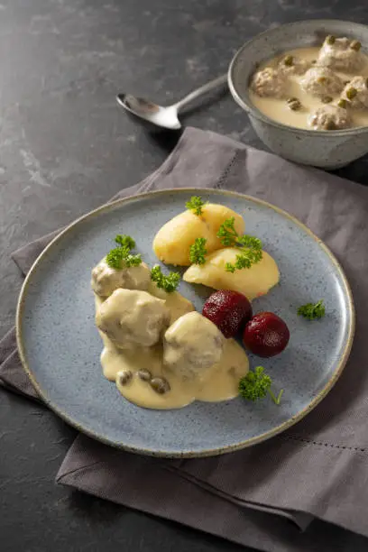 Koenigsberger Klopse or boiled meatballs in a white bechamel sauce with capers, potatoes and beetroot on a blue plate, dark gray background with napkin and copy space, selected focus, narrow depth of field