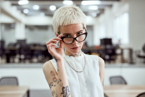 Portrait of young and confident blonde tattooed businesswoman adjusting her eyeglasses and looking at camera while standing in the modern working space. Business. Successful women