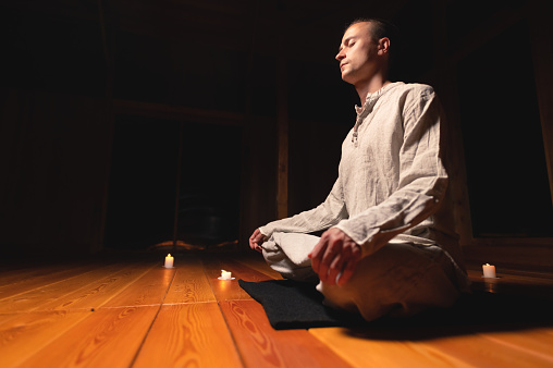 Attractive Caucasian young man meditates with closed eyes while sitting in lotus position in a dark room by candlelight