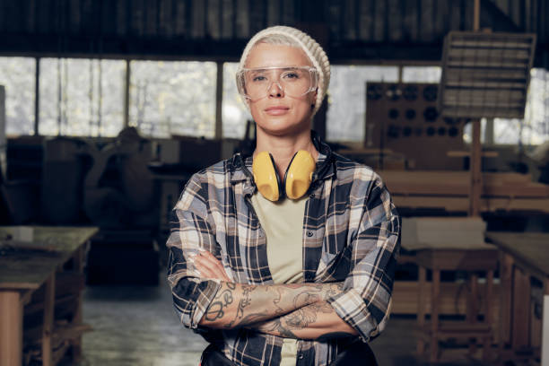 Portrait of female carpenter wearing protective eyewear Strong portrait of caucasian blond woman with hands crossed on chest, wearing protective gear, working as carpenter, standing at the factory. Masculine profession, gender equality, motivated woman concept carpenter stock pictures, royalty-free photos & images