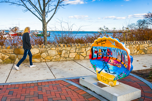 Plymouth, Massachusetts, USA- November 16, 2020-  A woman looks out over the fishing fleet in Plymouth Harbor as she walks past one of the several painted sea shells scattered through town to celebrate the 400th birthday of America's Home Town.