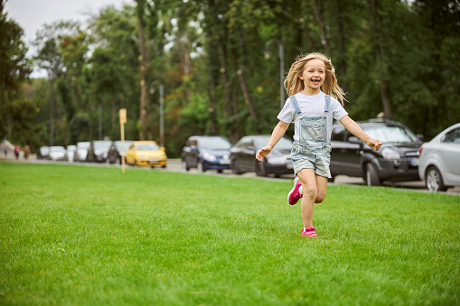 Portrait of beautiful girl in denim romper running in the city park on the green grass