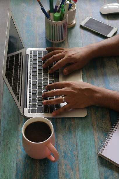 istock Image of unrecognisable person typing on a laptop computer at office work station, surrounded by coffee cup with black coffee, pen holder, potted cactus, wireless mouse, smart phone, note pad, pencils, pens, blue woodgrain background, focus on foreground 1286195232