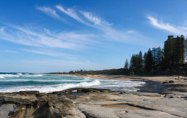 Beautiful Buddina Beach on the Sunshine Coast of Australia with beautiful turquoise water and unidentifiablee people down the beach and tall buildings and trees to the side and volcanic rocks in the foreground stock photo