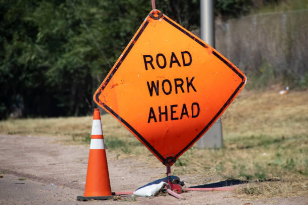 istock Battered Road Work Ahead sign and traffic cone sitting by road with blurred background 1286201893
