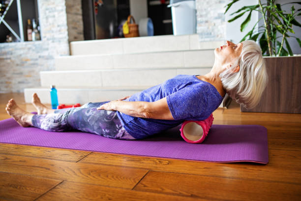Foam Roller Massage for Lower Back Pain senior woman stretching her back on a foam roller, lying down at on an exercise mat at home grey hair on floor stock pictures, royalty-free photos & images