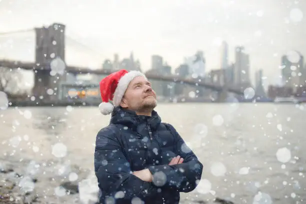 Photo of Mature man in a Santa Claus hat is walking during a snowfall near Brooklyn Bridge in New York on a snowy Christmas Eve. Surprised person admiring snowflakes. Winter Xmas holidays in NYC