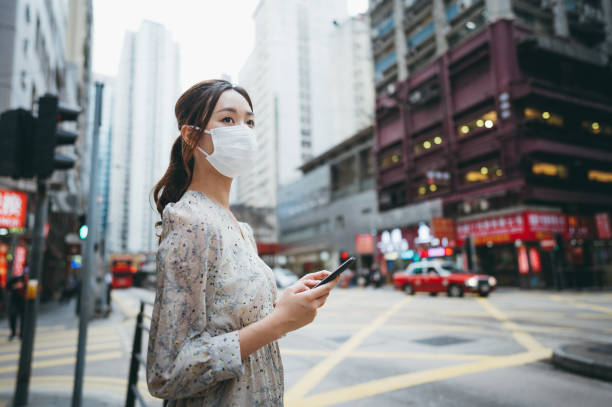 istock Asian young woman wearing a protective face mask, looking away while holding her smartphone on the street of Hong Kong. 1286214964