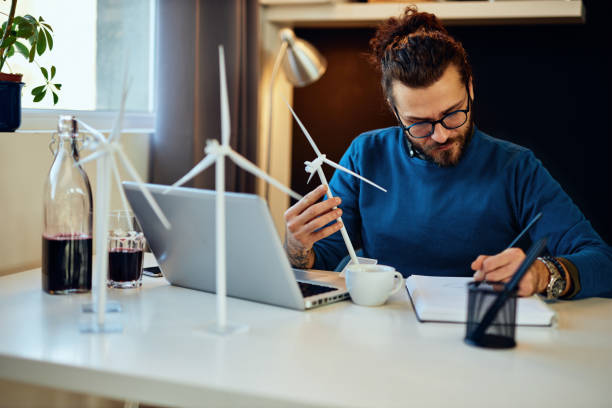istock Dedicated hardworking engineer sitting in his office, holding windmill model and drawing in notebook. Sustainable development concept. 1286225457