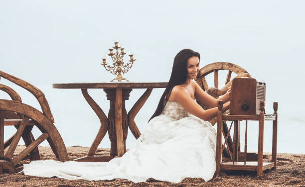 a linda noiva jovem em um vestido de noiva está esperando seu marido na praia passar momentos românticos, procurando música romântica na rádio nostálgica. - wedding dress audio - fotografias e filmes do acervo