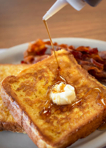 Two pieces of French Toast on a white plate with bacon blurred in the background. Two pieces of French Toast on a white plate with bacon blurred in the background.  Daub of butter melts on top while maple syrup is being poured.  Photo shot on location at diner. french toast bacon bread butter stock pictures, royalty-free photos & images