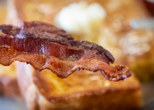 Close up view of a crispy slice of bacon with French toast blurred in the background. Close up view of a crispy slice of bacon with French toast blurred in the background. french toast bacon bread butter stock pictures, royalty-free photos & images