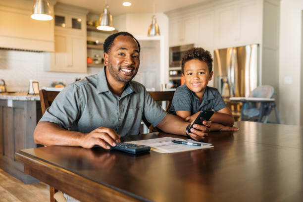 Father Teaching Son Home Finances A middle-aged man sits at his dining room table with his young son teaching him about home finances. He is working from home and telecommuting. financial education stock pictures, royalty-free photos & images