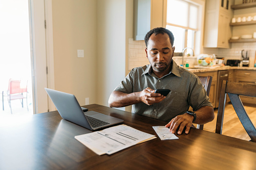 A middle-aged man sits at his dining room table with his paying his bills and banking using his mobile phone and laptop. He is taking a picture of his receipt for his online banking and tracking of expenses.