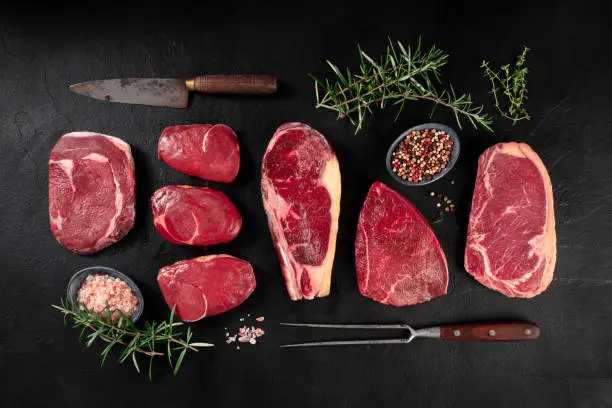 Photo of Meat, various cuts, overhead flat lay shot with a knife, a barbecue fork, salt, pepper, and rosemary