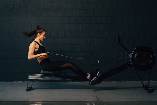 Side view of young muscular woman exercising in gym with rowing machine stock photo