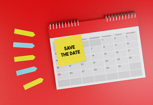 Desk calendar standing on red background. With Save The Date sticky note paper. Horizontal composition with copy space.