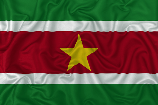 Suriname country flag on wavy silk textile fabric background.