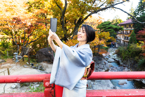 A senior Japanese woman dressed in Kimono enjoying the outdoors during the Autumn season in Japan with beautiful colored Maple leaves with her mobile phone taking a selfie.