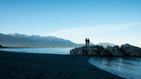 Couple standing on the rocks by the beach with snow-capped mountains in the distance, Kaikoura, New Zealand