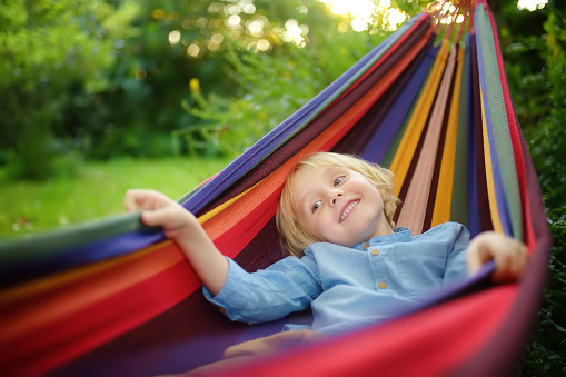 Cute little blond caucasian boy enjoy and having fun with multicolored hammock in backyard or outdoor playground. Summer outdoors active leisure for kids. Child relaxing and swinging in hammock.