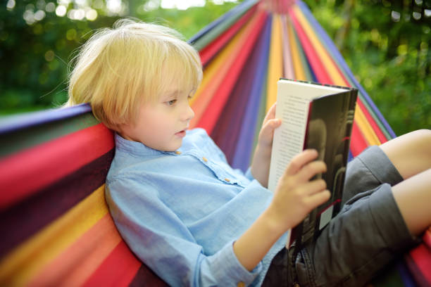 istock Cute little blond caucasian boy reading book and having fun with multicolored hammock in backyard or outdoor playground. Summer leisure for kids. Child swinging and relaxing in hammock. 1286287799