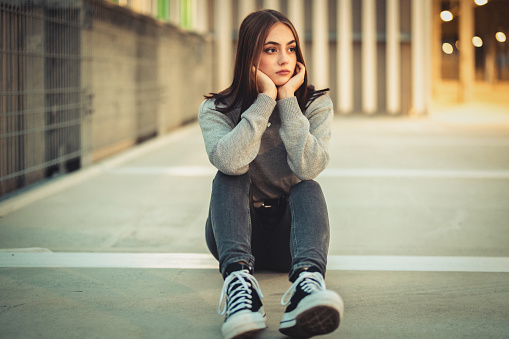 Sad and pensive looking lovesick teenage young woman having problems. Sitting alone on concrete floor of parking garage looking away, day dreaming. Young Millennial Generation Daily Life Concept.