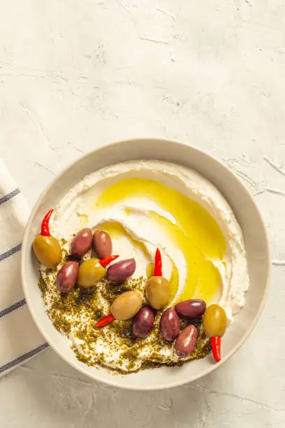 Photo of Popular middle eastern appetizer labneh or labaneh, soft white goat milk cheese with olive oil, hyssop or zaatar, olives on grey table, top view ,flat lay. Close up image.