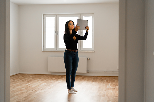 A young woman is standing in her new home with a digital tablet. She is using an augmented reality software to design and furnish her new empty apartment.