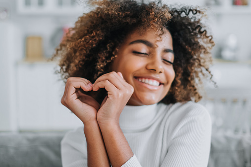 Cheerful model with perfect dark skin and white teeth hols hands in heart shape near face smiling widely at home close view