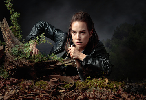 Portrait of a young woman with a knife. A serious girl in a black leather jacket with a knife climbs out of the pit in the forest.