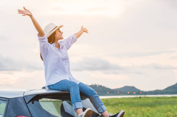 Happy Asian woman spread arms on car roof under sunset at seaside with nature background during travel in holiday. Confidence cheerful girl with dawn sky. Sun sea and mountain. Vacation concept feliz mujer asiática extendió los brazos en el techo del coche bajo la puesta del sol en la costa con fondo de la naturaleza durante el viaje en vacaciones. confianza chica alegre con el cielo del amanecer. mar de sol y montaña. concepto de vacaciones