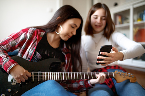 Two cheerful teenage girls at home, one is sitting on the floor and playing the guitar, her friend is sitting next to her and holding smart phone, they are socializing at home at time of COVID-19 lockdown