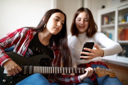 Two cheerful teenage girls at home, one is sitting on the floor and playing the guitar while looking at smart phone, her friend is sitting next to her and holding smart phone, they are socializing at home at time of COVID-19 lockdown