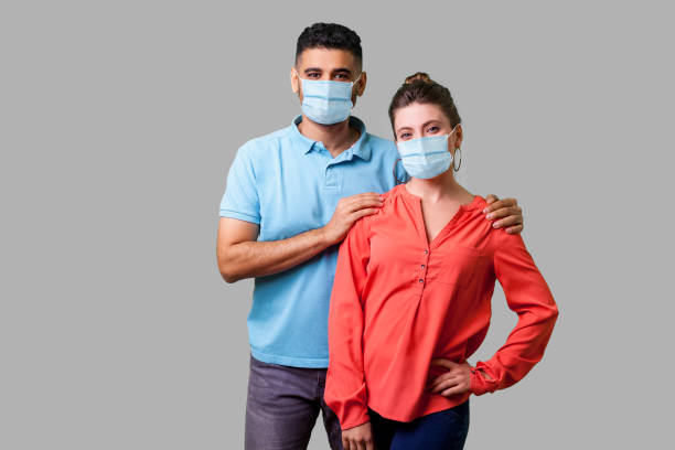 retrato de feliz pareja joven con máscara médica quirúrgica de pie juntos, mirando a la cámara con sonrisa dentada, hombre atento sosteniendo hombros femeninos. - friendly match illness cheerful head and shoulders fotografías e imágenes de stock