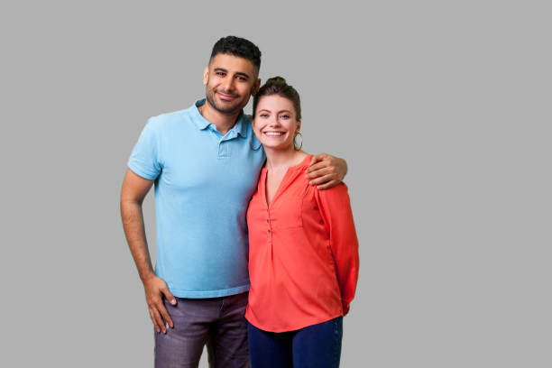 Portrait of young attractive family couple embracing. isolated on gray background, indoor studio shot Portrait of young attractive family couple in casual wear standing together, embracing and looking at camera with sincere smile, strong relations. isolated on gray background, indoor studio shot two parents stock pictures, royalty-free photos & images