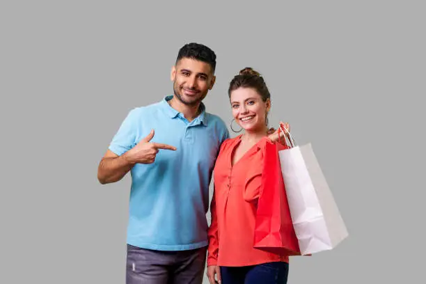 Photo of Satisfied shoppers. Portrait of happy bearded young man pointing at shopping bags. isolated on gray background