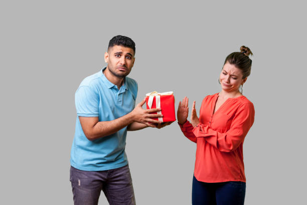 Portrait of upset resentful man in casual clothes giving present to naughty beautiful woman. isolated on gray background Portrait of upset resentful man in casual clothes giving present to naughty beautiful woman, dissatisfied girl showing refusal gesture, bad gift. isolated on gray background, indoor studio shot unliked stock pictures, royalty-free photos & images