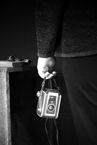 Black and white photograph of a woman's hand holding a generic vintage camera in front of a dark background and wooden crate.