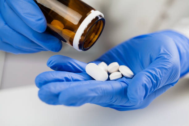 Mmedical practitioner wearing blue surgical latex gloves holding medicine bottle UK NHS Doctor, nurse or GP medical practitioner wearing blue surgical latex gloves holding medicine bottle, taking out several white pills, COVID-19 corona virus disease epidemic, Coronavirus global pandemic antiviral drug stock pictures, royalty-free photos & images