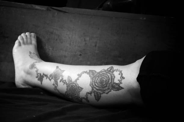 Black and white photograph of a woman's flower tattoo on the leg in front of a dark background and wooden crate.