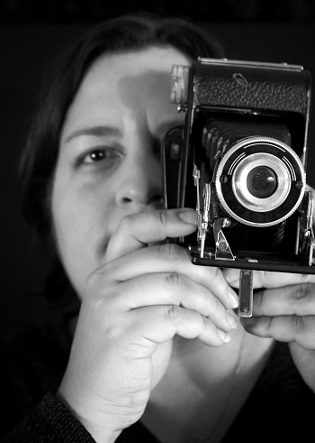 Black and white studio shot photograph of a woman taking a picture with an old generic camera in front of a dark background.