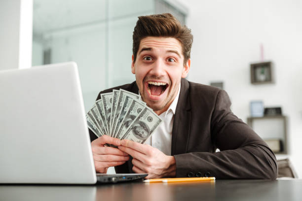 istock Excited young businessman showing money using laptop computer. 1286341001