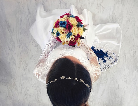 Upper view of a bride standing up, holding her bouquet, before her wedding. Her dress is white with blue details in it, matching the colours of the flowers on her bouquet.