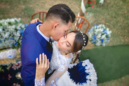 A beautiful young Brazilian newlywed couple hugging each other in tenderness, with some flowers decorating the floor. The bride looks tranquil as she holds her husband. Her left hand has a gold ring on the finger.